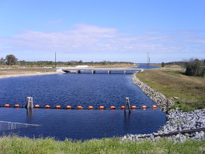 [A wide shallow stream with a line of orange bouys across it in the foreground and a bridge in the middle ground. There are stones lining both sides of the water inlet.]